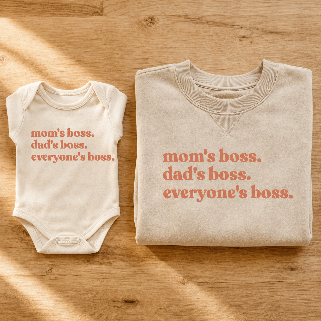 WeeBoss 'mom's boss. dad's boss. everyone's boss.' baby bodysuit and adult sweatshirt flat lay on warm honey wood — peach serif text on cream and sand cotton, coordinated not matching