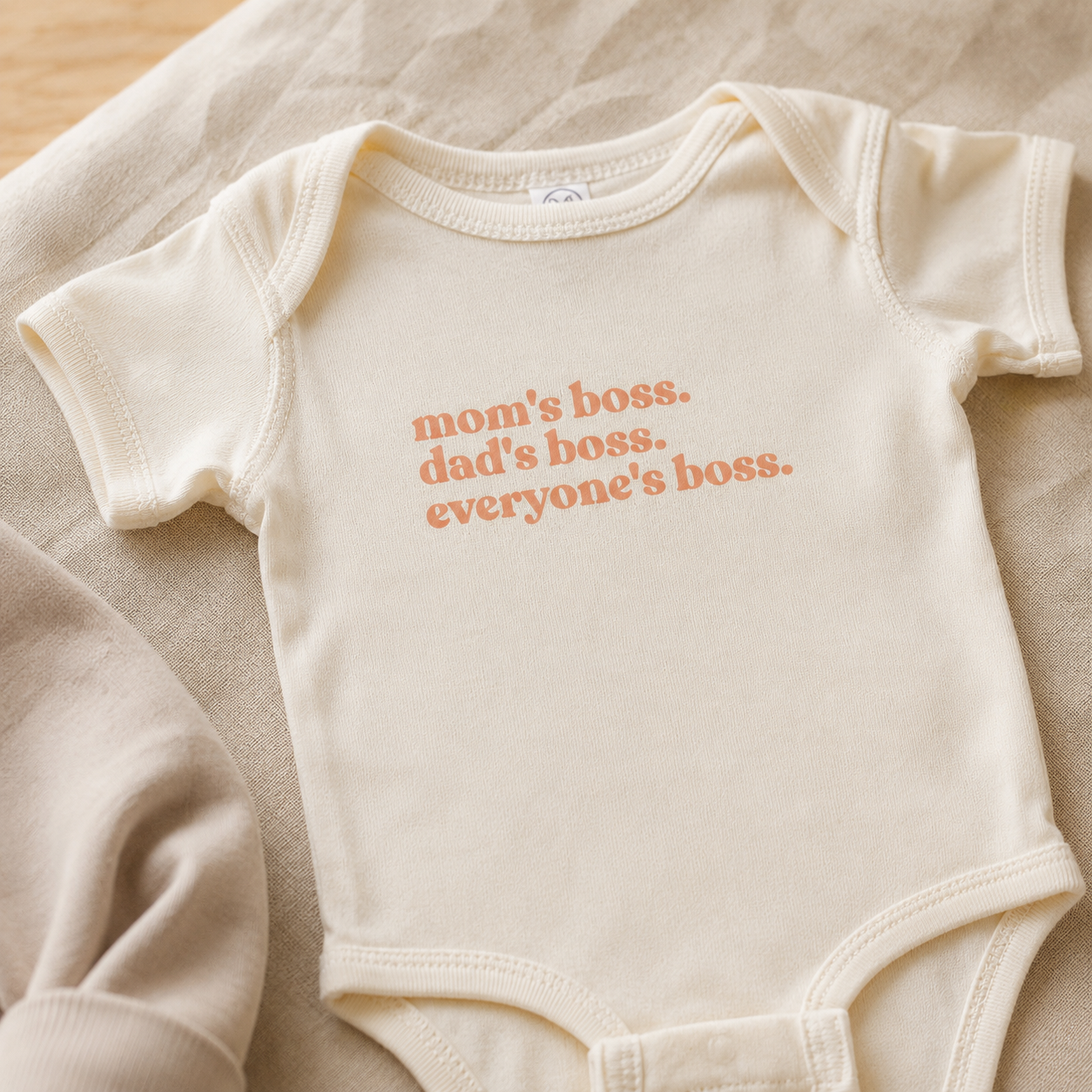WeeBoss baby bodysuit close-up on soft oat fabric — 'mom's boss. dad's boss. everyone's boss.' in peach serif text, natural cream cotton, warm diffused light