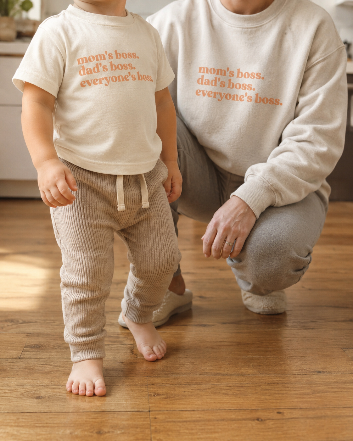 Toddler and parent wearing coordinated WeeBoss 'mom's boss. dad's boss. everyone's boss.' tee and sweatshirt — cream and warm sand, warm wood floor, bright home kitchen in background