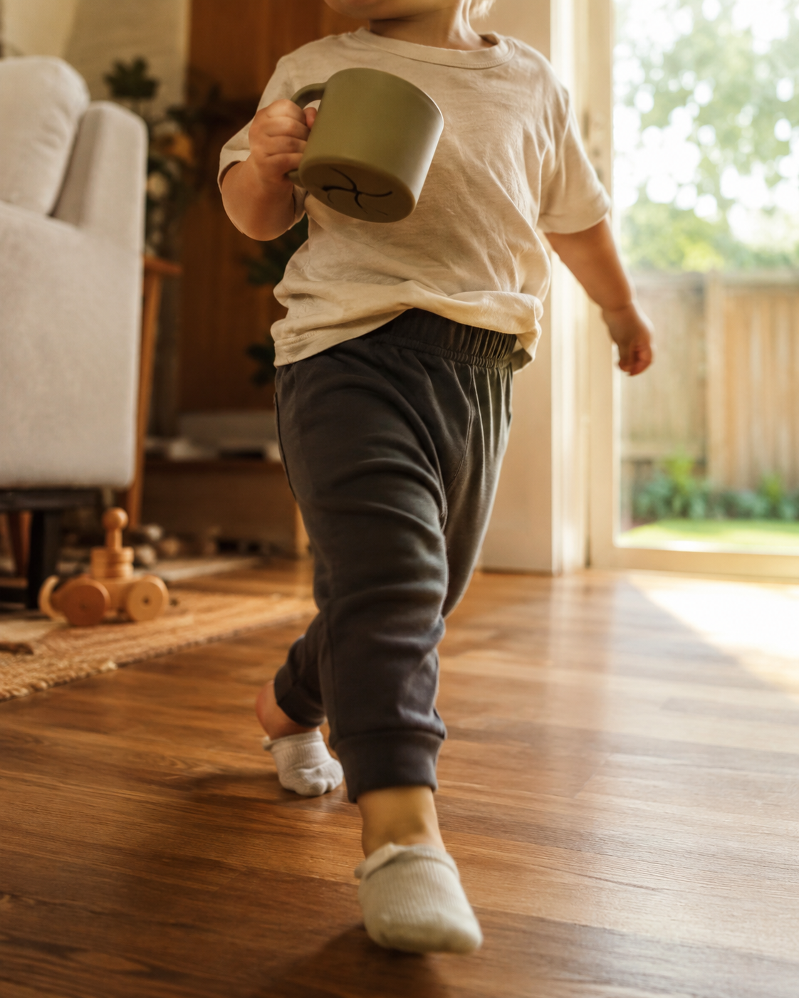 Toddler torso and legs mid-movement — soft tee, comfy pull-on pants, one sock slightly slipping, snack cup in hand. Real clothes for a kid who knows where they are going.